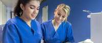 Zwei Pflegefachfrauen im blauen Kasack am Patientenwagen.