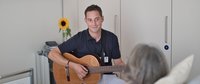 Musiktherapeut mit Gitarre am Bett einer Patientin.
