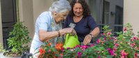 Ergotherapeutin unterstützt Patientin, die im Innenhof das Blumenbeet gießt.