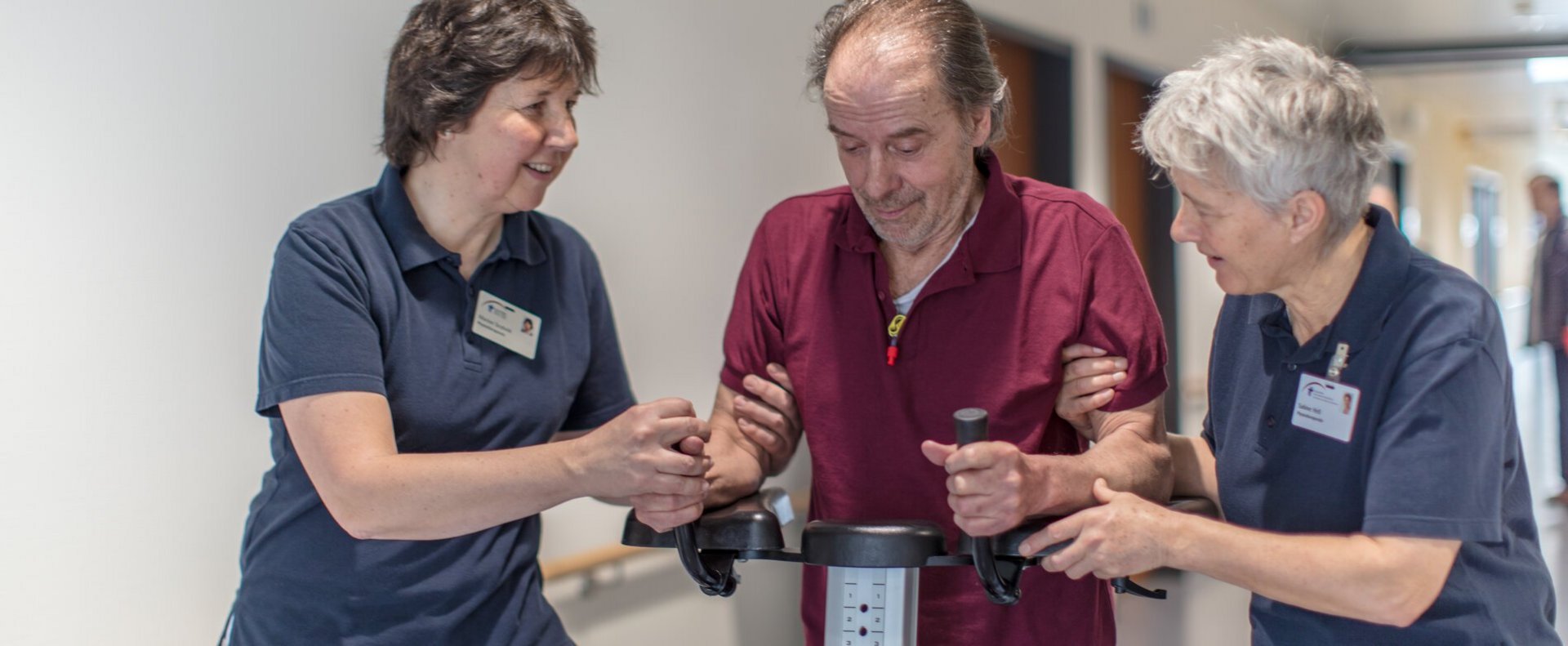 Zwei Physiotherapeutinnen unterstützen Patienten am Gehwagen.