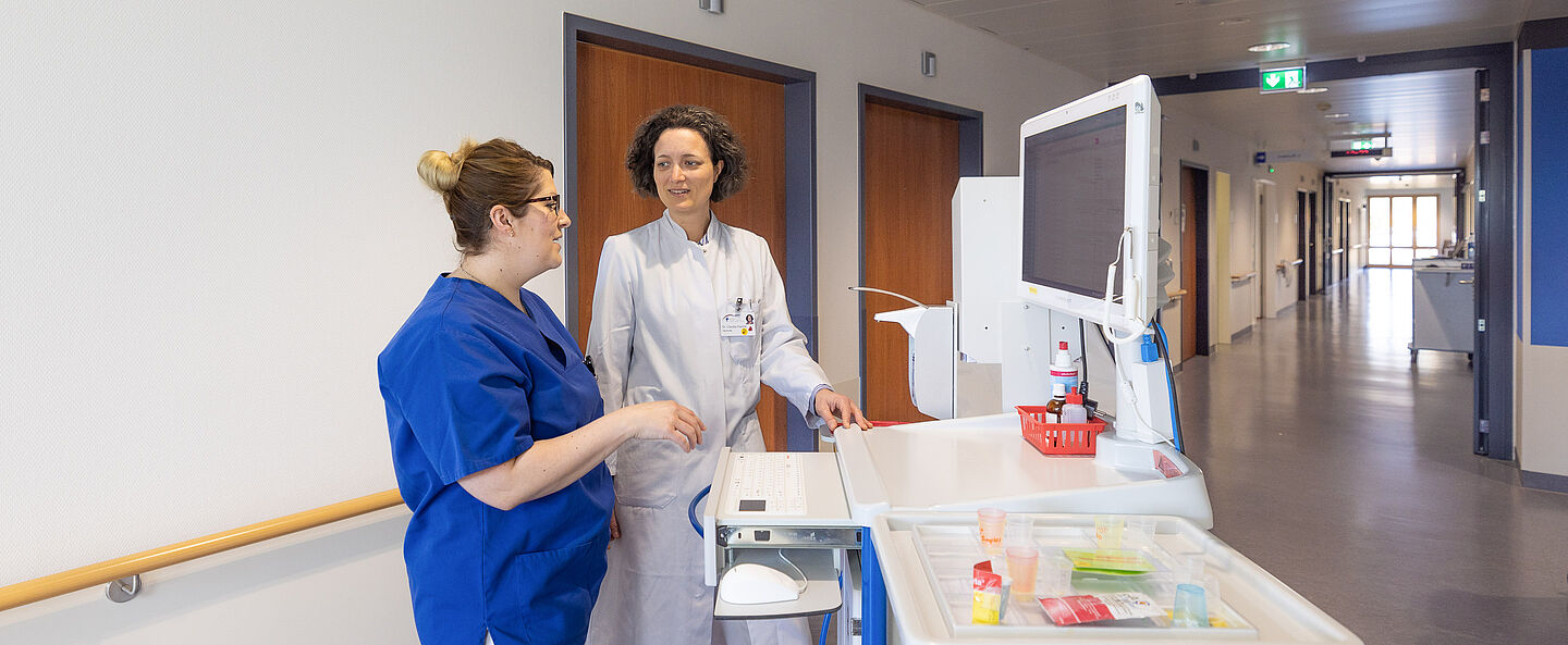Ärztin und Pflegefachfrau im Gespräch am Patientenwagen.